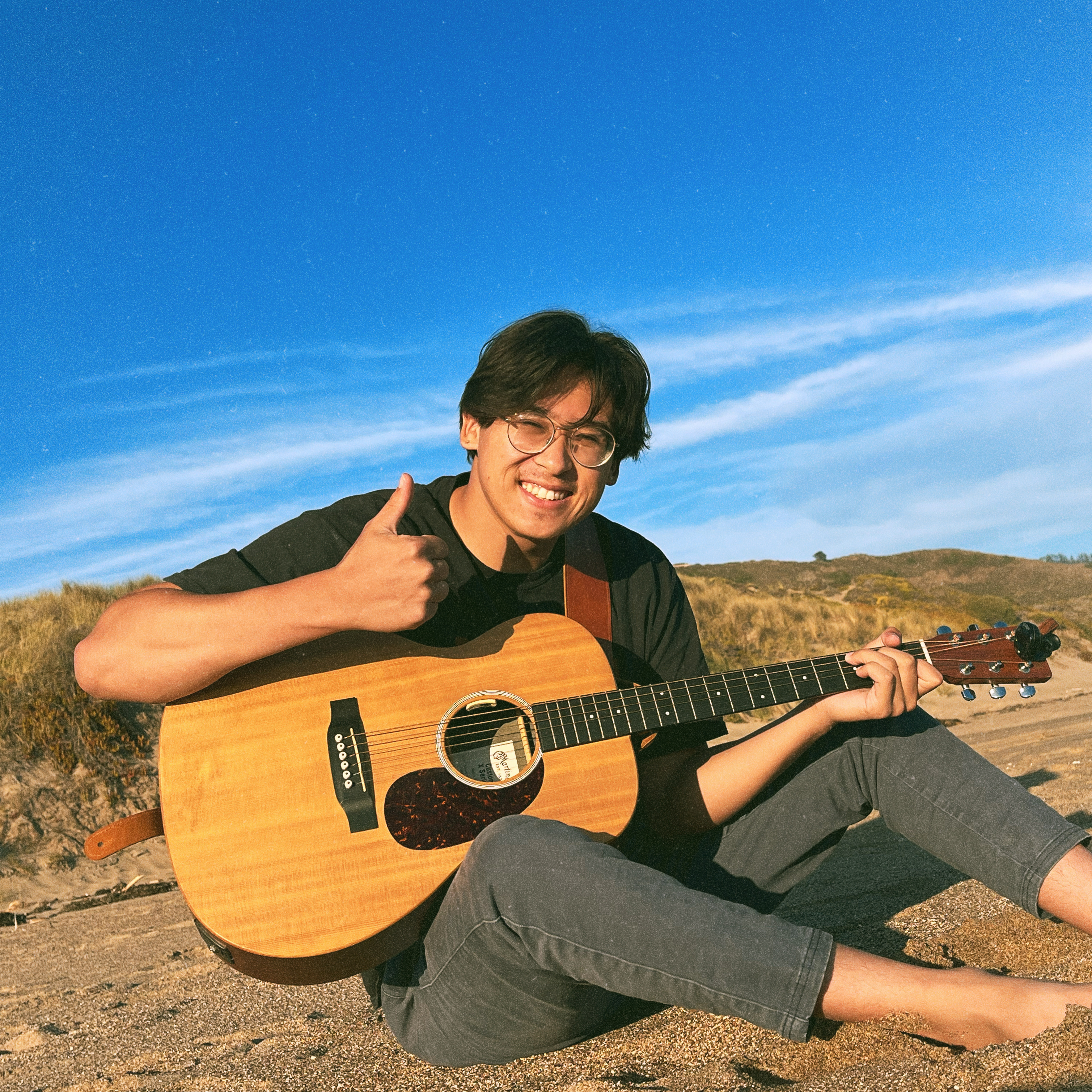 Noah smiling with an acoustic guitar on coastal dunes